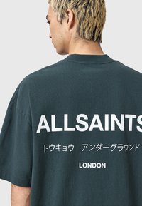 Jeune personne avec des cheveux blonds courts portant un t-shirt oversize vert foncé sur lequel est écrit ALLSAINTS, texte japonais et LONDON au dos.