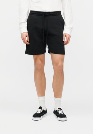 Vīrietis, kurš valkā melnas šorti ar aukliņu, baltu garām piedurknēm kreklu, baltas zeķes un melnas kurpes ar šņorēm, stāv pret tukšu fonu.