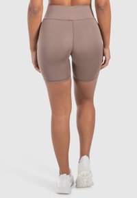 Pantaloni corti da ciclismo marroni a vita media realizzati in tessuto elasticizzato, con una texture liscia e un design aderente, indossati con sneaker bianche.
