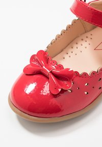 Zapato de charol rojo con puntera redonda, que presenta un gran lazo decorativo y detalles perforados en los laterales. Incluye una correa ajustable.