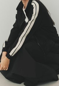 Chaqueta negra con cremallera hecha de una tela suave, que presenta largas franjas blancas a lo largo de las mangas. El diseño incluye puños y un cuello de punto.