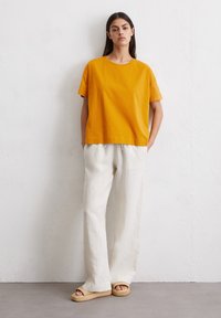 Geel katoenen t-shirt met korte mouwen, gecombineerd met roomkleurige linnen broek met wijde pijpen en natuurlijke geweven plateausandalen. Eenvoudig ontwerp en relaxed fit.