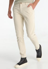 Chinos en coton beige avec une coupe ajustée, dotés de poches avant et d'un bouton marron à la taille. Associé à des baskets montantes noires.