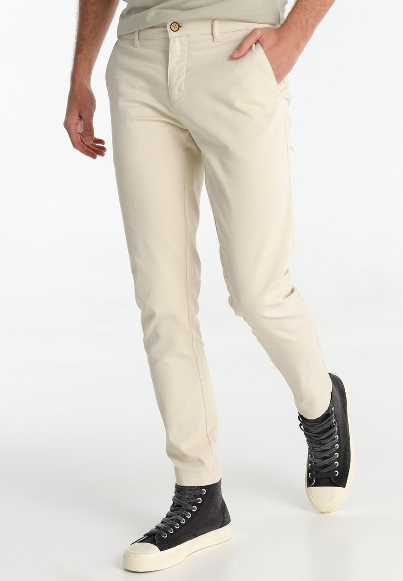 Chinos en coton beige avec une coupe ajustée, dotés de poches avant et d'un bouton marron à la taille. Associé à des baskets montantes noires.