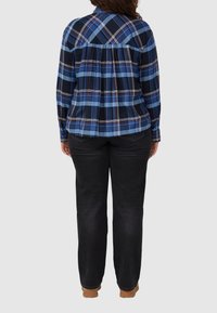 Person standing facing away wearing a blue plaid long-sleeve shirt, black jeans, and brown shoes on a light gray background.