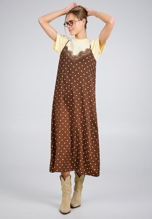 Kvinde med briller iført en brun slipkjole med polkaprikker over en gul t-shirt og beige støvler, som justerer sit hår med begge hænder.