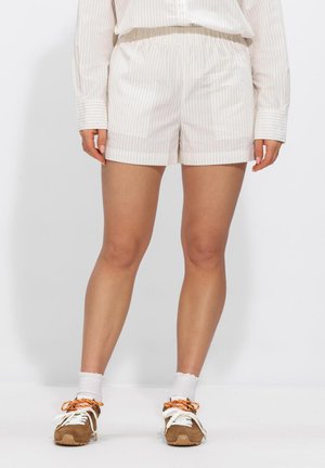 Femme portant un short beige à fines rayures, une chemise à manches longues assortie, des chaussettes blanches et des baskets marron, debout devant un fond blanc.