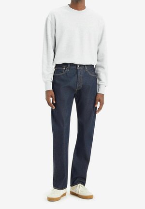 Persoon draagt een lichtgrijs sweatshirt, donkerblauwe rechtvallende spijkerbroek en witte sneakers met rubberen zolen, staand tegen een effen achtergrond.