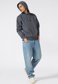 Grijze hooded sweatshirt met logo, voorvak en lange mouwen, gecombineerd met lichtblauwe, loszittende jeans en witte sneakers.