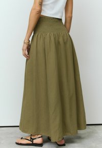 Jupe maxi kaki avec taille smockée, en tissu fluide et à texture subtile, associée à des sandales plates noires.