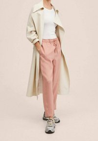 Beige trenchcoat, witte tanktop, roze broek met trekkingkoord en grijze sneakers. Het trenchcoat heeft brede revers en de broek heeft een relaxed fit.