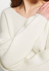 Femme portant un pull en maille côtelée de couleur crème avec un large col, levant un bras et touchant son épaule avec l'autre main.