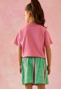T-shirt en coton rose avec une coupe décontractée, associé à une jupe verte présentant des rayures verticales roses et bleues, se tenant devant un fond rose.