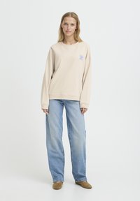 Sweat-shirt beige à manches longues avec broderie florale bleue, assorti à un jean large bleu clair et des chaussures à enfiler en daim marron.