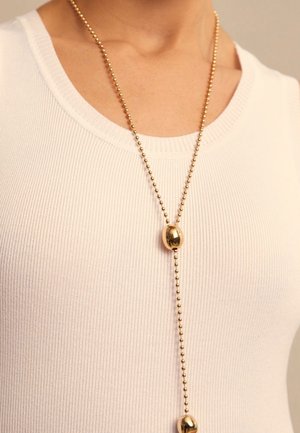 Collier en perles dorées avec des accents ovales, suspendu à une fine chaîne, assorti à un débardeur blanc texturé à côtes.