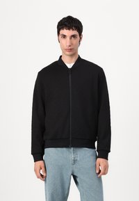 Svart zip-up bomberjakke med teksturert mønster, ribbede mansjetter og søm, kombinert med lyseblå jeans.