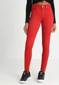 Mujer vestida con pantalones rojos ajustados, top negro de manga larga corta y zapatillas Fila en blanco y negro, de pie contra un fondo neutro.