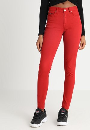Mujer vestida con pantalones rojos ajustados, top negro de manga larga corta y zapatillas Fila en blanco y negro, de pie contra un fondo neutro.