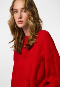 Red knitted sweater with a textured diamond pattern, featuring a relaxed fit and ribbed neckline, made of soft, warm wool blend.
