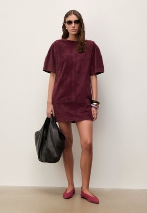 Donna che indossa un miniabito bordeaux a maniche corte, ballerine abbinate, occhiali da sole, porta una grande borsa in pelle nera e indossa braccialetti a un polso.