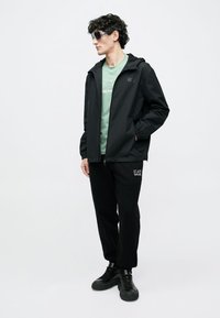 Mustaa kevyttä hupparia, johon yhdistetty vihreä t-paita ja mustat housut. Päällä on tummat kengät ja aurinkolasit, neutraalissa taustassa.