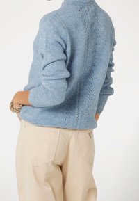 Persoon in een lichtblauwe gebreide trui met een patroon op de rug, gecombineerd met beige broeken en een gouden kettingarmband.