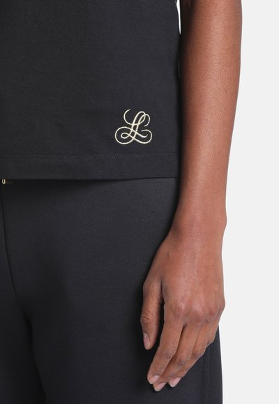 Gros plan sur la main d'une personne à côté d'une chemise et d'un pantalon noirs avec une petite lettre manuscrite brodée claire "L" sur l’ourlet de la chemise.