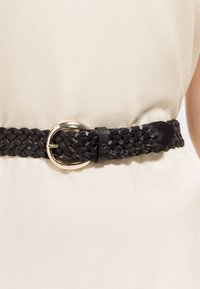 Cintura in pelle nera intrecciata con fibbia rotonda dorata allacciata intorno a una vita rivestita di tessuto beige.