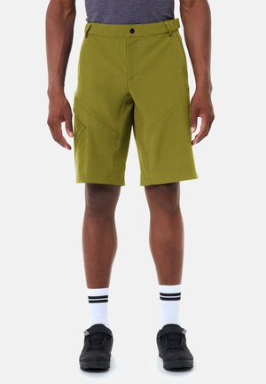 Uomo che indossa pantaloncini oliva al ginocchio, calzini bianchi con strisce nere e scarpe sportive nere, in piedi contro uno sfondo semplice.