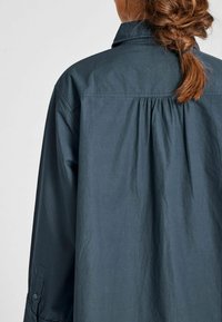 Donna con i capelli castani intrecciati, che indossa una maglietta lunga a maniche lunghe grigia scura e mostrata di spalle su uno sfondo chiaro semplice.