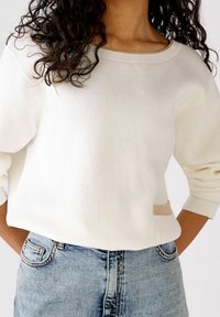 Biały sweter dziany z szerokim dekoltem i oversized rękawami. Zawiera beżowy akcent po boku. Połączony z jasnoniebieskimi dżinsami.