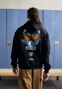Čierna mikina s grafickým potlačou orla a mesiaca na chrbte. Obsahuje kapucňu na šnúrku a rebrované manžety. Neformálny štýl.