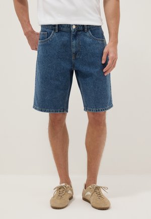 Hombre con pantalones cortos de mezclilla azul de longitud media, camisa blanca y zapatillas beige, de pie con las manos relajadas a los lados.