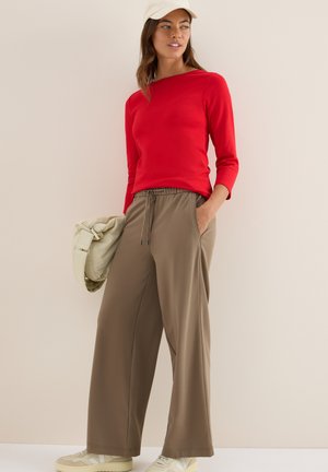 Maglietta rossa a maniche lunghe abbinata a pantaloni beige a gamba larga. I dettagli notevoli includono una cintura elastica e tasche. Scarpe beige e una borsa chiara.