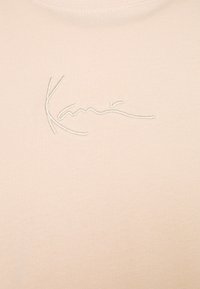 Lys beige t-skjorte med en myk og jevn tekstur; har brodert kursiv tekst "Kani" i en litt kontrasterende trådfarge.
