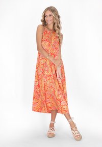 Ein lebhaftes orangefarbenes Maxikleid mit kunstvollen Paisleymustern, dünnen Trägern und einem fließenden Design, kombiniert mit hellen, riemenförmigen Wedge-Sandalen.