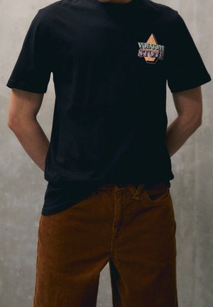 Personne portant un t-shirt noir avec le logo Volcom Stone et un pantalon en velours côtelé marron, debout les mains derrière le dos devant un fond gris.