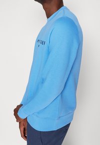 Felpa azzurra chiara con collo a giro, polsini e orlo a coste. Presenta un logo stampato nero sul davanti. Materiale in cotone morbido.