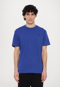 Μπλε βαμβακερό μπλουζάκι με κοντά μανίκια, στρογγυλή λαιμόκοψη και διακριτικό μαύρο λογότυπο στη δεξιά πλευρά του στήθους, σχεδιασμένο για άνετη εφαρμογή.