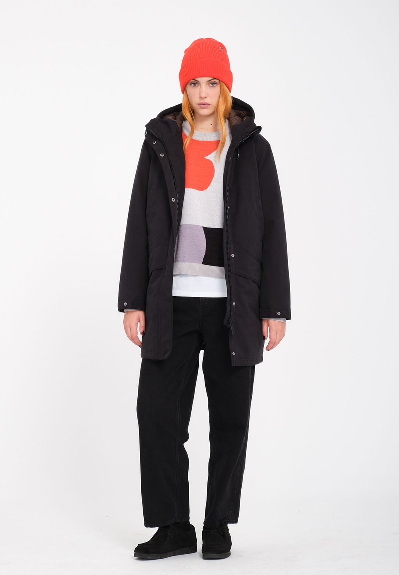 Parka nera con cappuccio, maglione grigio e colorato sotto, pantaloni neri e berretto lavorato a maglia arancione. Calzature nere.