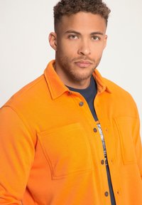 Camisa chaqueta de color naranja brillante con botones a presión, dos bolsillos frontales y un collar, hecha de un material texturizado. Llevada sobre una camiseta de color marino.