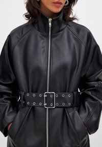 Svart fusk läderjacka med hög krage, framdragkedja och bred bälte med silveröglor. Lång lös passform med sidfickor.