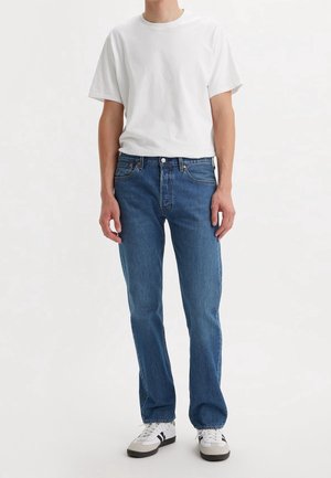 Personne portant un t-shirt blanc uni à manches courtes, un jean bleu droit et des baskets blanches avec des rayures noires, debout sur fond blanc.