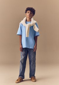 Plava majica kratkih rukava, kremasti šal omotan oko vrata, sive traperice, smeđe cipele s vezicama i teksturiranim đonom, stojeći na neutralnoj pozadini.