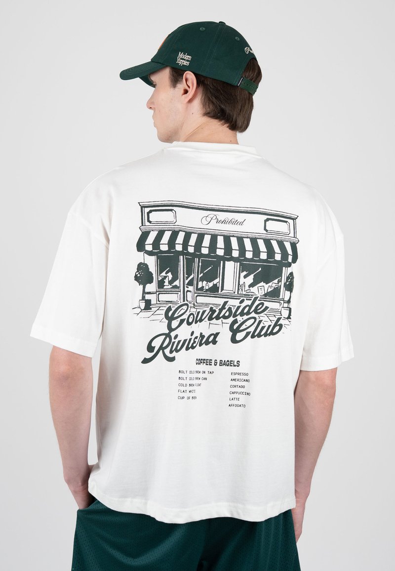 Homme portant un t-shirt blanc oversize avec un graphisme de vitrine de café noir et le texte « Courtside Riviera Club » ainsi qu'un menu, casquette vert foncé et short.