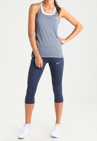 Šedý sportovní nátělník s bílými doplňky, doplněný tmavě modrými tříčtvrtečními legínami. Modelka má na sobě bílé tenisky. Hladká látka, přiléhavý střih.