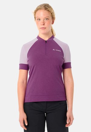 Frau mit hell lockigem Haar trägt ein violettes und lavendelfarbenes kurzärmeliges Radtrikot mit Frontreißverschluss und schwarze Hose.