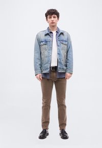 Νεαρός με σγουρά μαλλιά που φοράει τζιν μπουφάν πάνω από καρό πουκάμισο και λευκό μπλουζάκι, καφέ παντελόνι, μαύρα παπούτσια, στέκεται μπροστά σε απλό φωτεινό φόντο.