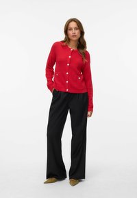Une femme se tient debout, portant un cardigan tricoté rouge à boutons, un pantalon taille large noir et des chaussures à talons pointus vert olive, devant un fond blanc uni.