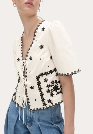 Hvid bluse med sort blomsterbroderi, bølget kanter, puffede ærmer og binding foran, båret med blå højttaljede jeans.